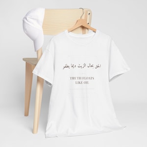 May include: White t-shirt with the text "TRUTH FLOATS LIKE OIL" in English and Arabic script. A white baseball cap rests on a wooden chair. The shirt hangs on a wooden hanger.