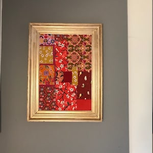 Peut inclure: Une œuvre d'art en patchwork encadrée avec un cadre doré. Le patchwork est composé de différents tissus dans des tons de rouge, orange, jaune et vert. Les tissus ont des motifs différents, notamment floraux, géométriques et paisley.