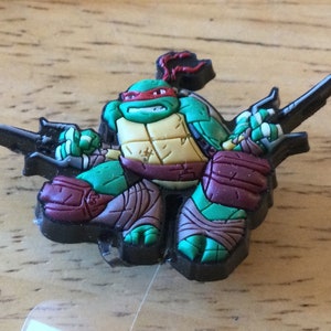 May include: A green Teenage Mutant Ninja Turtles character with a red mask and brown and purple clothing holding two black katanas.