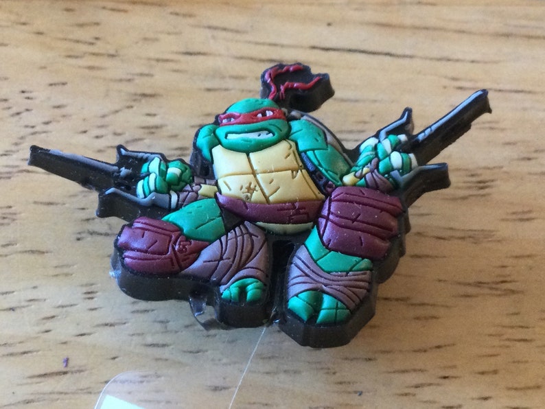 May include: A green Teenage Mutant Ninja Turtles character with a red mask and brown and purple clothing holding two black katanas.