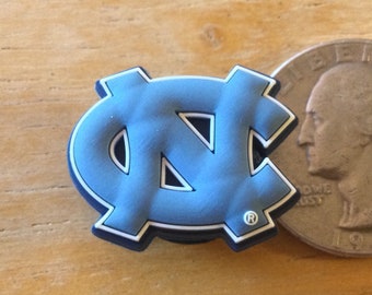 May include: A blue and white UNC Tar Heels logo charm. The logo is a stylized "NC" with a white outline and a registered trademark symbol.
