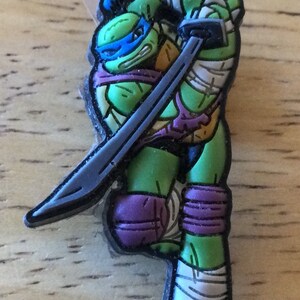 May include: A green Teenage Mutant Ninja Turtle action figure with a purple bandana and a silver katana sword. The figure is wearing white bandages on its arms and legs.