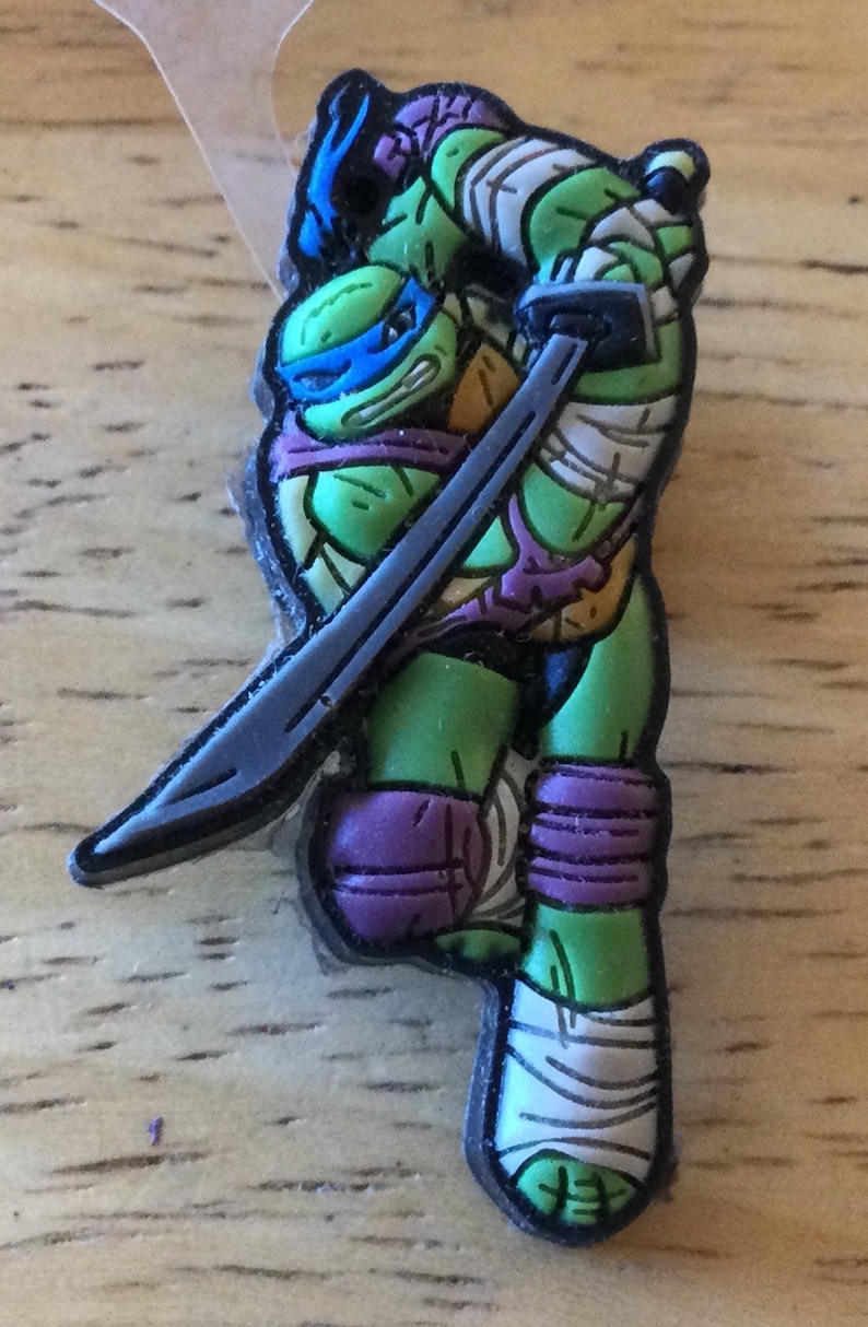 May include: A green Teenage Mutant Ninja Turtle action figure with a purple bandana and a silver katana sword. The figure is wearing white bandages on its arms and legs.