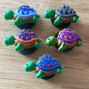 Può includere: Cinque ciondoli per scarpe a forma di tartaruga colorati con gusci verdi e dettagli arancioni. Le tartarughe hanno gusci di diversi colori: blu, viola, grigio e due tonalità di blu.