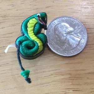 May include: A green and yellow cobra snake charm with a black cord and a green bead. The charm is about the size of a quarter.