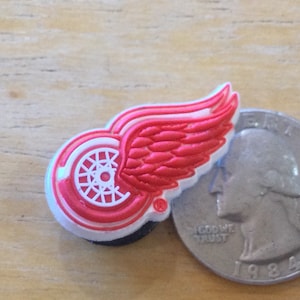 May include: A red and white emblem featuring a winged wheel design, likely a team logo. The emblem is placed next to a quarter coin for size comparison. The logo has a white outline and a black base.