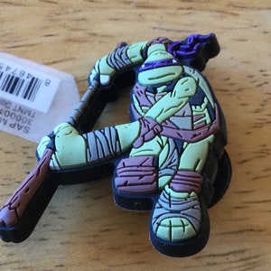 May include: A green, black and purple Teenage Mutant Ninja Turtles character keychain. The character is holding a brown wooden stick. The keychain is made of rubber.