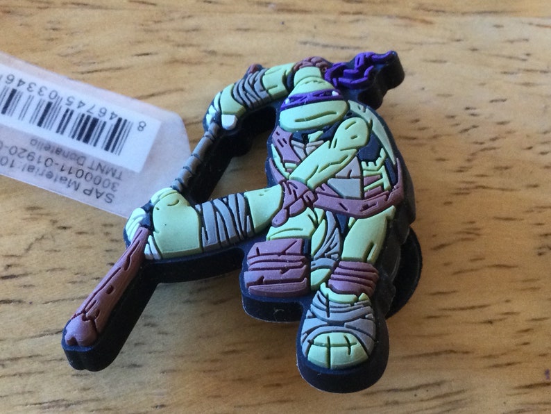 May include: A green, black and purple Teenage Mutant Ninja Turtles character keychain. The character is holding a brown wooden stick. The keychain is made of rubber.