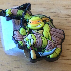 May include: A green Teenage Mutant Ninja Turtles character with an orange mask and brown bandana, holding a katana and a sai. The figure is made of rubber and has a black chain attached to it.