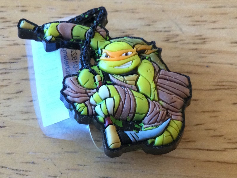 May include: A green Teenage Mutant Ninja Turtles character with an orange mask and brown bandana, holding a katana and a sai. The figure is made of rubber and has a black chain attached to it.