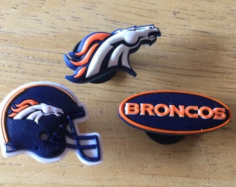 May include: Three Denver Broncos shoe charms. One charm features the team's logo, a white horse with an orange mane and tail. Another charm features a blue and white football helmet with the team's logo. The third charm is a blue oval with the word "BRONCOS" in white letters.