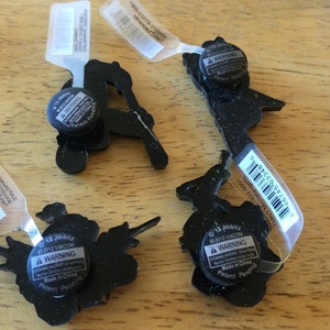 May include: Four black and green Jibbitz shoe charms with warning labels. The charms feature a variety of characters, including a turtle, a ninja turtle, a skateboarder, and a witch. The labels state "&copy;2012 VIACOM" and "A WARNING CHOKING HAZARD - Small Parts. Not for Children Under 3 Years. Made in China. Patent Pending."