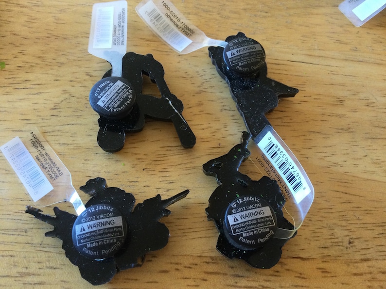 May include: Four black and green Jibbitz shoe charms with warning labels. The charms feature a variety of characters, including a turtle, a ninja turtle, a skateboarder, and a witch. The labels state "&copy;2012 VIACOM" and "A WARNING CHOKING HAZARD - Small Parts. Not for Children Under 3 Years. Made in China. Patent Pending."