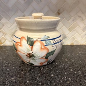 May include: A cream-colored ceramic jar with a lid, featuring a hand-painted floral design in shades of orange, white, and green. The jar has blue stripes and the words "God is Faithful" written on the side. The jar is sitting on a dark countertop.