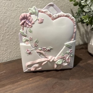 May include: A ceramic heart-shaped picture frame in the form of an envelope. The frame is white with pink and lavender accents, featuring floral and ribbon details. The frame is approximately 6 inches tall.