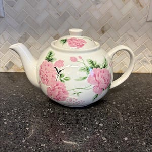 May include: White ceramic teapot with a lid and handle, adorned with pink floral patterns and green leaves. The teapot is set against a dark countertop and a light-colored tile backsplash. The teapot is designed for brewing tea.