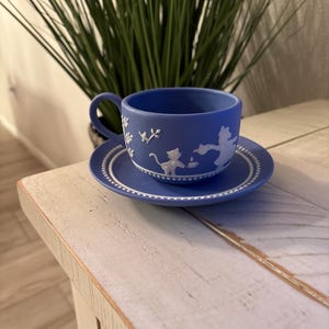 May include: A blue teacup and saucer set with white relief designs of cats and floral patterns. The cup has a handle and sits on a matching saucer. The set is placed on a white and light brown wooden table.