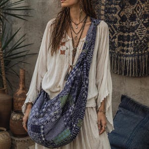 May include: A long-sleeved off-white blouse with a loose fit and a subtle embroidered detail on the chest, paired with a large, slouchy crossbody bag made from patchwork fabric in shades of navy blue, white, and green with geometric patterns.