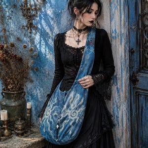 May include: A blue denim hobo bag featuring a large, light-colored sun graphic with swirling rays. The bag has a wide shoulder strap and a drawstring closure with a silver-toned metal ring and a small white bead accent at the bottom center.