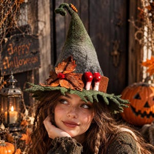 May include: A dark green felted pointed hat featuring a curled tip with a small orange caterpillar, a brown band, two red and white mushrooms, and a cluster of brown autumn leaves attached to the front with a small red berry accent.