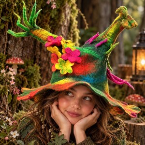 May include: A felted hat featuring a tall, pointed crown with green, yellow, and red blended colors. The hat is decorated with several bright pink and yellow felted flower appliqués and includes long, thin, branch-like extensions protruding from the top.