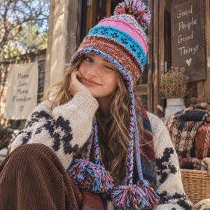Könnte beinhalten: Eine Strickmütze mit einem mehrfarbigen Muster aus rosa, blauen und braunen horizontalen Streifen und einem Bommel oben. Die Mütze hat zwei lange, geflochtene seitliche Zöpfe, die in großen, mehrfarbigen Wollbommeln an den Seiten enden.