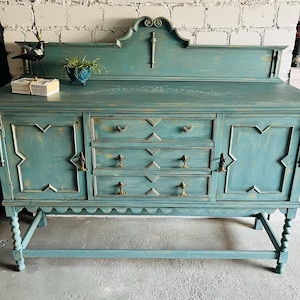 Könnte beinhalten: Ein türkisfarbenes antikes Sideboard mit drei Schubladen und zwei Schranktüren. Das Sideboard hat aufwendige Details, darunter dekorative Beschläge und geschnitzte Akzente. Die Oberseite hat eine dekorative Rückseite und der Text "BRIGHTON'S PATISSERIE" ist sichtbar.