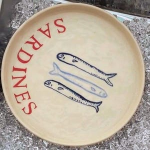 Puede incluir: Un plato de cerámica color crema con la palabra "SARDINES" en letras rojas. Tres peces azules están pintados en el plato. El plato está sobre un lecho de hielo.