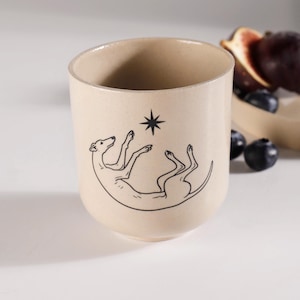 May include: A beige ceramic mug featuring a black line art illustration of a greyhound dog lying on its back with its paws reaching toward a small black star symbol centered above the dog. The mug has a smooth, matte finish and a cylindrical shape.