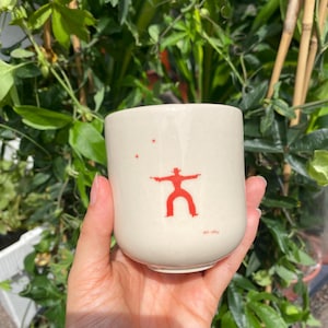 Puede incluir: Taza de cerámica color crema con un gráfico de vaquero rojo y pequeñas estrellas rojas. La taza es cilíndrica y parece hecha a mano. La taza se sostiene en una mano, con follaje verde en el fondo.