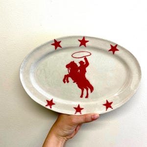 Puede incluir: Plato ovalado de cerámica con base crema y un gráfico rojo de vaquero y caballo en el centro. Estrellas rojas espaciadas alrededor del borde. El plato es sostenido por una persona. Aproximadamente 30 cm de largo.