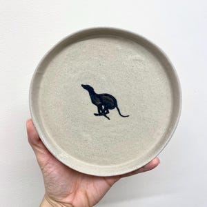 以下が含まれることがあります： オフホワイトの丸い陶器の皿で、走っている動物の濃い青色のイラストが描かれています。 皿にはわずかに盛り上がった縁と斑点のあるテクスチャがあります。 動物のイラストは皿の中央に配置されています。