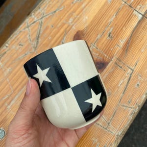 May include: A ceramic cup with a black and white chequered pattern, featuring white star designs. The cup has a rounded shape and is held in a hand. The cup is sitting on a wooden surface.