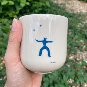 May include: A cream-colored ceramic mug featuring a blue graphic of a cowboy silhouette holding two pistols with two small blue stars above the figure and the text YEE-HAW printed in small blue letters near the bottom of the mug's exterior.