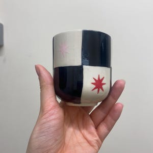 May include: A ceramic cup with a geometric design. The cup is divided into black and cream-coloured squares. Pink and red starburst designs are on the cream-coloured squares. The cup is held in a hand.