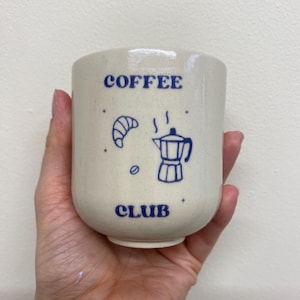 May include: A cream-colored ceramic coffee mug with the words "COFFEE CLUB" in blue. The mug features blue illustrations of a croissant, coffee bean, and a coffee pot. The mug is held in a hand.