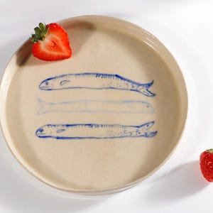 May include: A beige ceramic plate with a blue fish design. The plate features three blue fish illustrations. Two fresh strawberries are placed on the plate. The plate is round with a slightly raised edge.