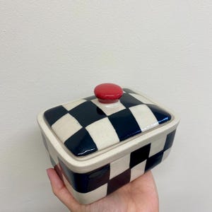 May include: A rectangular ceramic butter dish with a lid. The dish and lid are decorated with a black and white checkerboard pattern. The lid has a small, round, red knob on top. The dish is held in a hand.