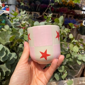 Op de afbeelding: Een roze en crème keramische beker met een geruit patroon en rode sterren. De beker wordt in een hand gehouden. De achtergrond is wazig en toont een bloemenwinkel met diverse planten en bloemen. De beker is ongeveer 8 cm hoog.
