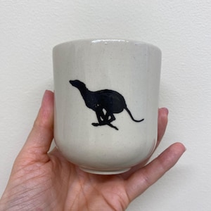 May include: A cream-colored ceramic cup with a black silhouette of a running dog. The cup has a rounded shape and a speckled texture. The dog illustration is on the front of the cup.