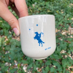 May include: A cream-colored ceramic cup with a speckled finish featuring a blue graphic of a person riding a rearing horse, two small four-pointed stars above the rider, and the text "yee-HAW" printed in blue near the bottom of the cup.