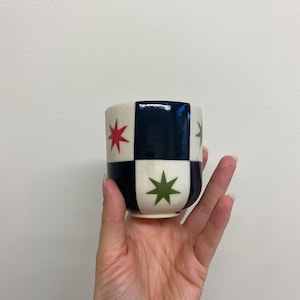 May include: A small ceramic cup with a checkered pattern of navy blue and cream squares. Red and green star designs are featured on the cream squares. The cup is held in a hand against a plain white background.
