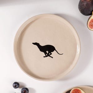 Op de afbeelding: Een rond, beige keramieken bord met in het midden een zwart silhouet van een rennende windhond. Het bord heeft een gladde, matte afwerking en een licht opstaande rand. Het ontwerp is eenvoudig en focust op het contrast tussen de donkere hondengrafiek en het lichte, neutrale oppervlak van het servies.