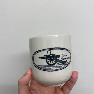 May include: Cream ceramic mug with a black oval design depicting a cannon and the words "The Gunners." The mug has a cylindrical shape and a smooth, glossy finish. A hand is holding the mug.