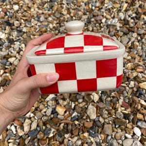 May include: A rectangular ceramic butter dish featuring a vibrant red and off-white checkered pattern. The dish includes a matching lid with a small, rounded knob handle on top. The design consists of alternating solid red and cream-colored squares.