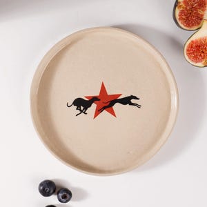 May include: A round, off-white ceramic plate featuring a central graphic of a red five-pointed star flanked by two black silhouettes of running greyhounds. The plate is accompanied by two sliced fig halves and three fresh blueberries.