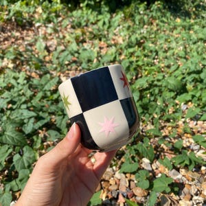 May include: A ceramic mug featuring a black and cream checkered pattern. The design is accented with small, multi-coloured star shapes in shades of pink, lime green, and red, distributed across the alternating squares of the mug's exterior surface.