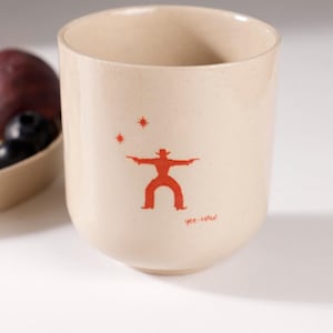 Peut inclure: Un mug en céramique beige orné d'un motif rouge représentant un cow-boy tenant deux pistolets, surmonté de deux petites étoiles. Le texte "yee-HAW" est inscrit en rouge sous la silhouette. Le mug présente une finition mouchetée.