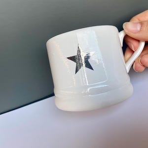 May include: White ceramic mug with a black star design. The mug has a rounded base and a curved handle. The star is centered on the front of the mug.