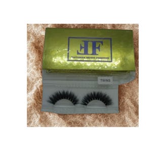 May include: A pair of black false eyelashes labeled "Twins" in a clear plastic case. The case is inside a gold box with a blue logo and the text "Flattering Beauty Cosmetics".